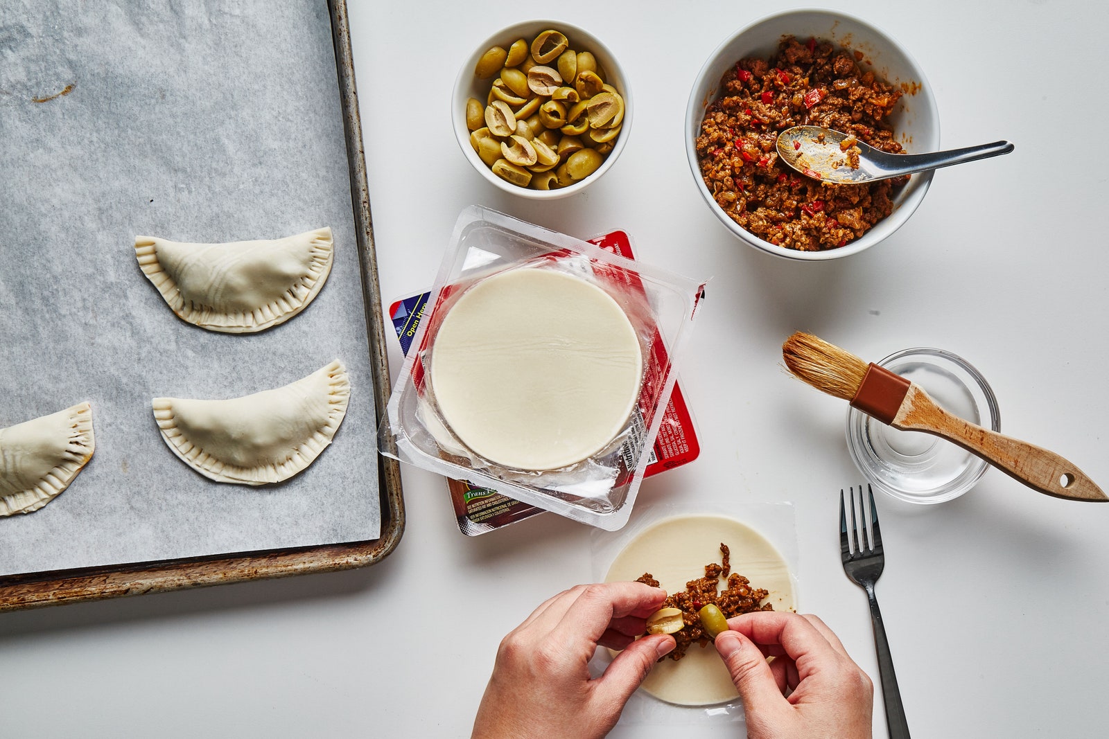 Hands adding olives to beef empanada filling.