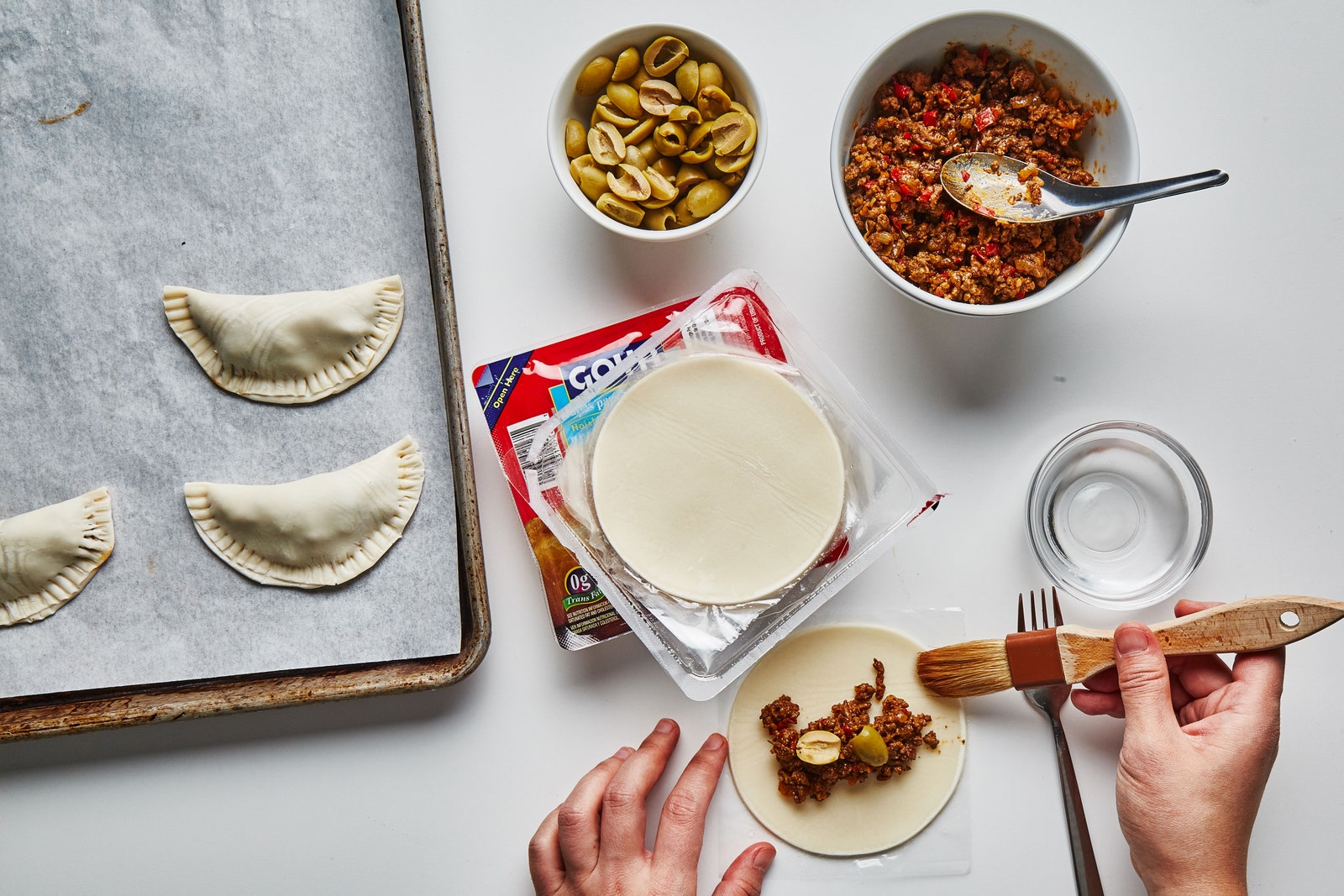 Hands brushing water around edge of beef empanada dough.