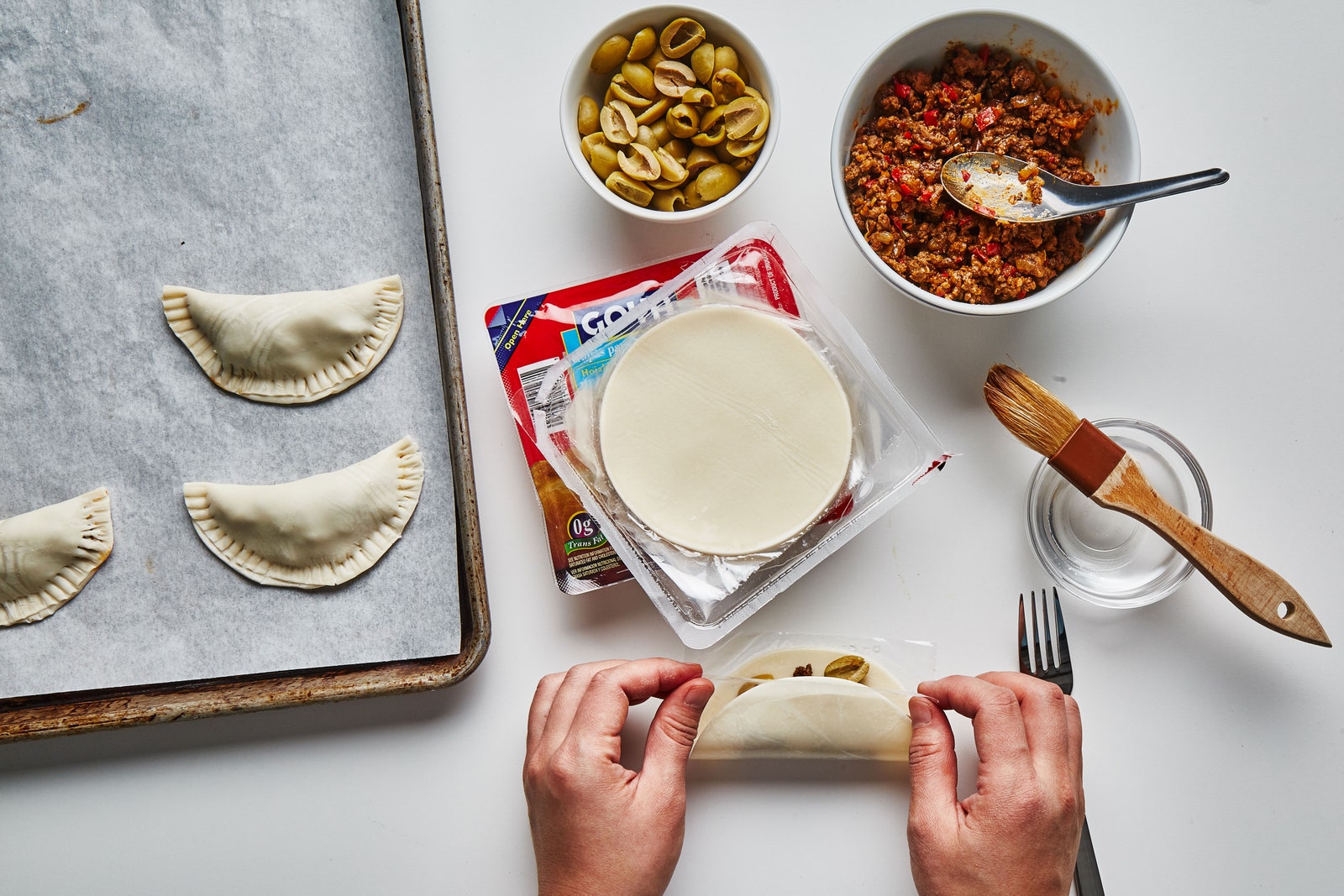 Hands folding empanada dough around a beef picadillo filling.