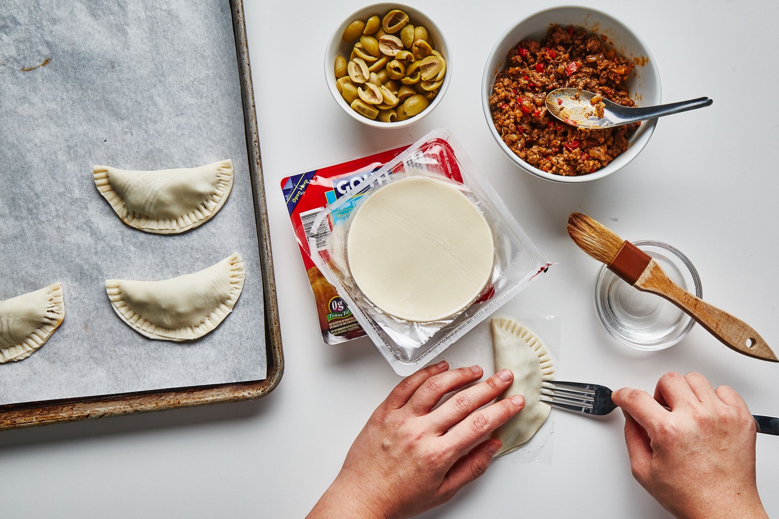 Hands using a fork to crimp the rounded edge of a beef empanada.